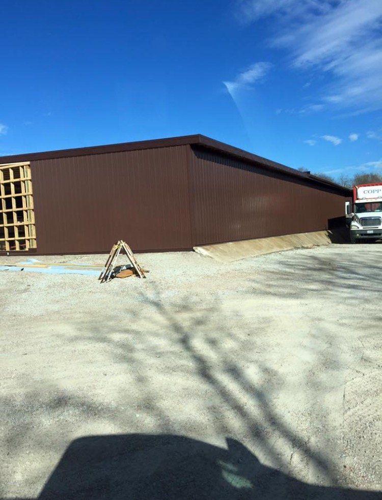 Brown warehouse on a gravel lot under a blue sky, with a truck parked at right and a barricade in front.