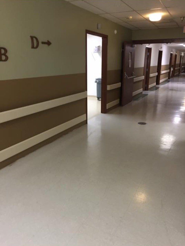 Hospital hallway with beige walls, white floor, and open doorways labeled with room letters.