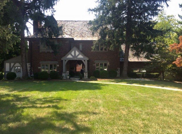 Brick house with white trim and large front lawn, framed by tall trees under bright sunlight