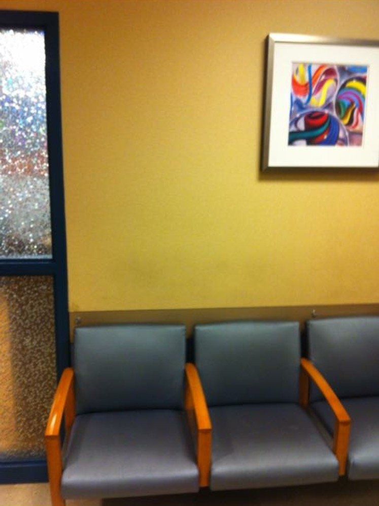 Waiting area with three gray chairs against a yellow wall and a colorful framed print above them