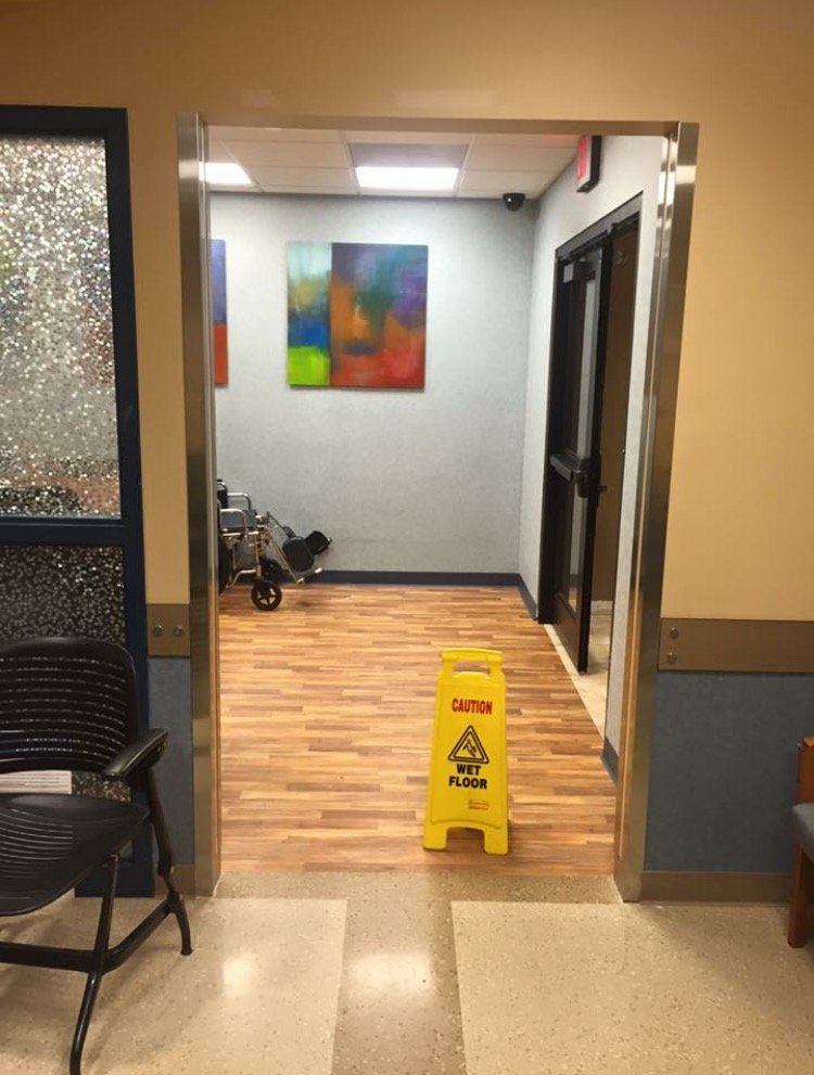 Hallway with yellow wet floor sign, black chair, colorful wall art, and doorway to a gray room.