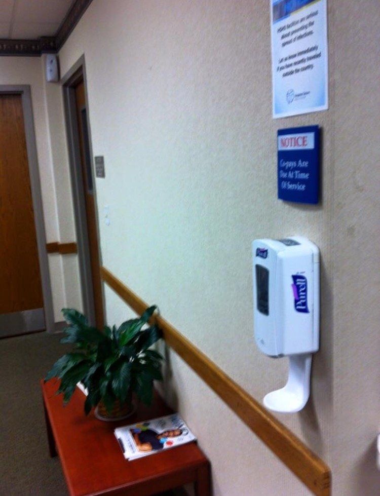 Hallway wall with hand sanitizer dispenser, notice signs, and a small table with a plant and magazines