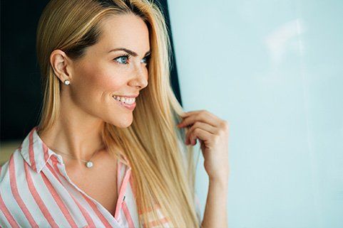 A woman with long blonde hair is smiling and touching her hair.