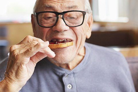 An elderly man wearing glasses is eating a piece of bread with jam on it.