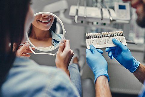 A woman is looking at her teeth in a mirror while a dentist holds a tooth color chart.