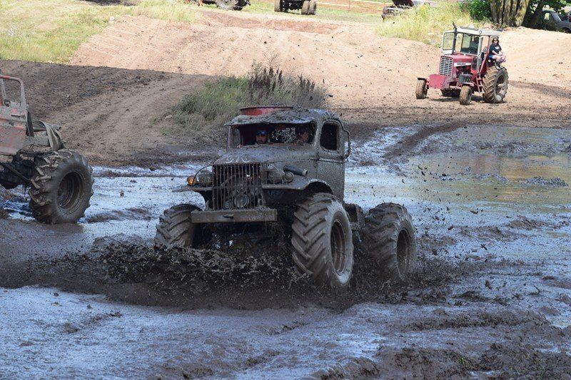 Mud-covered off-road trucks racing through a muddy track, with a red vehicle in the background