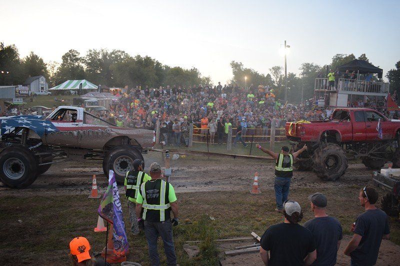 Monster trucks facing off on a dirt track before a crowded grandstand at sunset