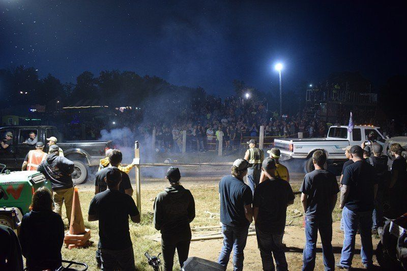 Crowd gathered at a nighttime outdoor event near smoke, floodlights, and parked vehicles