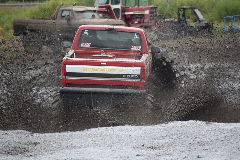 Red off-road truck splashing through muddy water on a dirt track, with another truck in the background