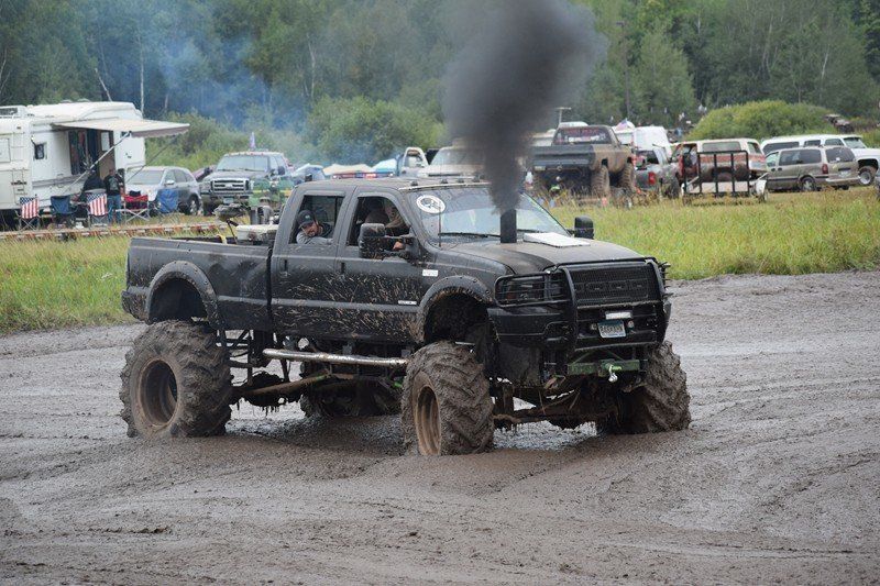 Black lifted pickup truck splashing through mud at a muddy outdoor truck event, with smoke rising from the hood