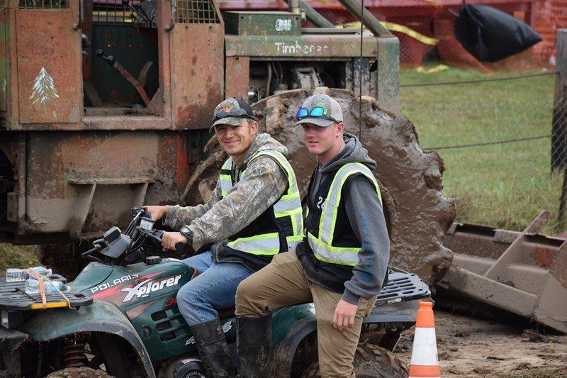 Two men in safety vests sit on an ATV beside construction equipment and orange cones outdoors