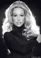 Black-and-white portrait of a woman with wavy blonde hair in a dark turtleneck, looking at the camera.