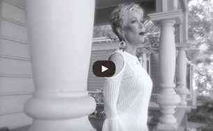A black and white photo of a woman in a white dress standing on a porch.