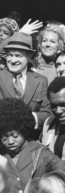 Crowd of smiling people, including a man in a hat at center, gathered outdoors.