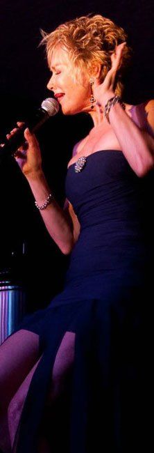 Singer in a navy dress performing on stage with a microphone, lit by purple-pink lights.