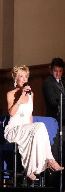Woman in a white dress speaking into a microphone onstage, with a man standing behind her.