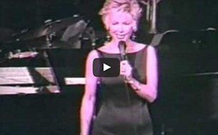 Singer in a black dress holding a microphone on a dimly lit stage.