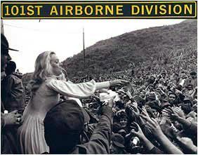 Woman reaches toward a crowd below a “101st Airborne Division” banner on a hillside.