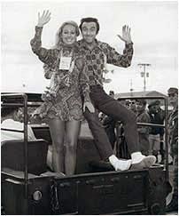 Two people waving while sitting on the back of a jeep at an outdoor event