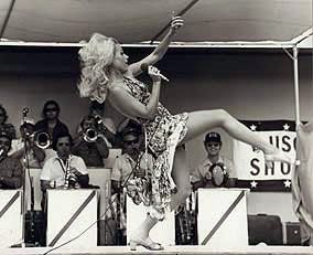 Black-and-white stage performance with a singer in a patterned dress striking a dramatic pose before a band