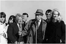 Group of people standing outdoors, including a man in a hat and a blonde woman, posing for a photo.