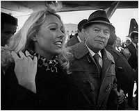 Black-and-white photo of a smiling woman beside a man in a hat at a crowded outdoor event