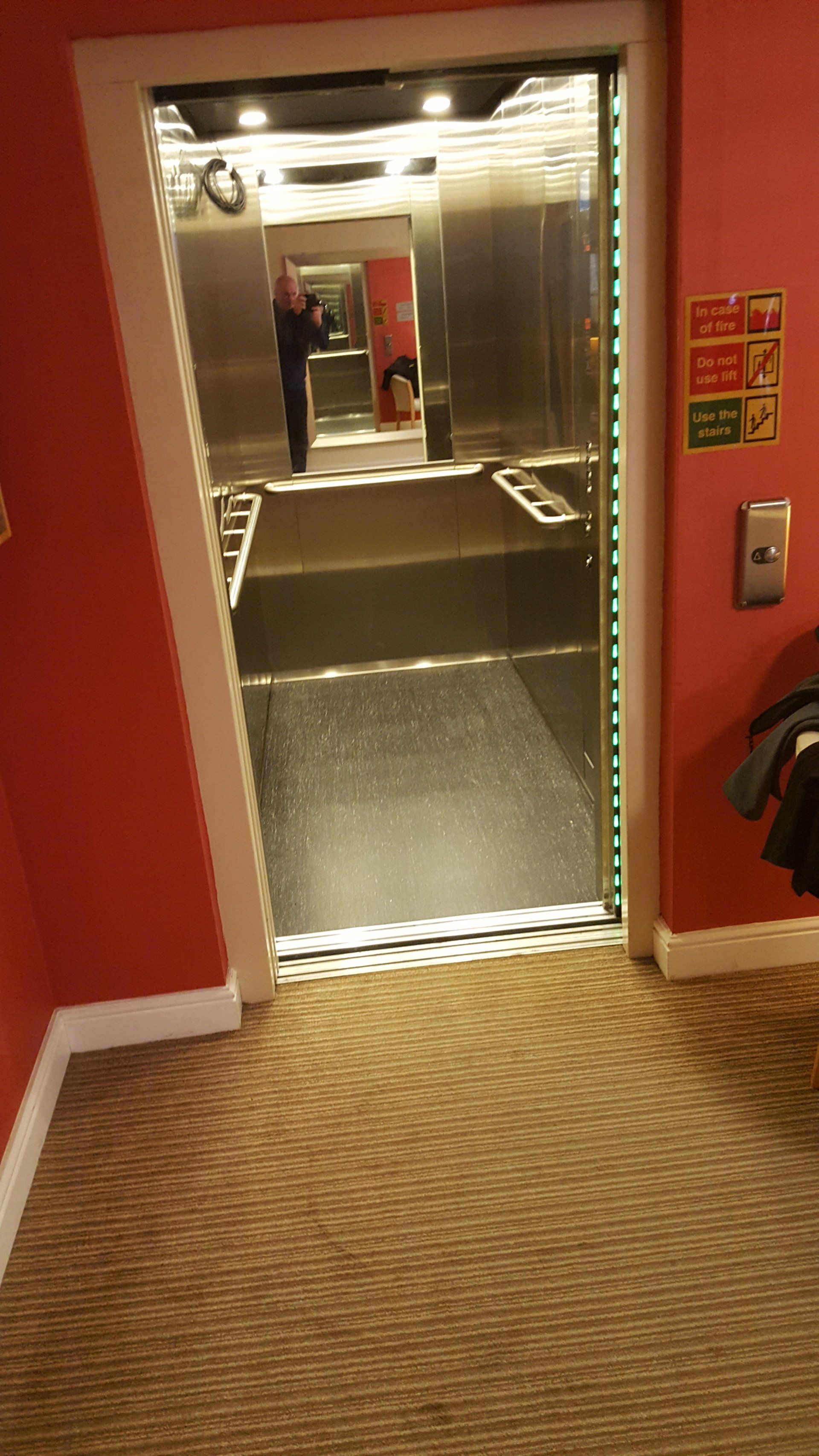 Elevator with open doors in a red-walled hallway; a person is reflected in the elevator's mirrors.