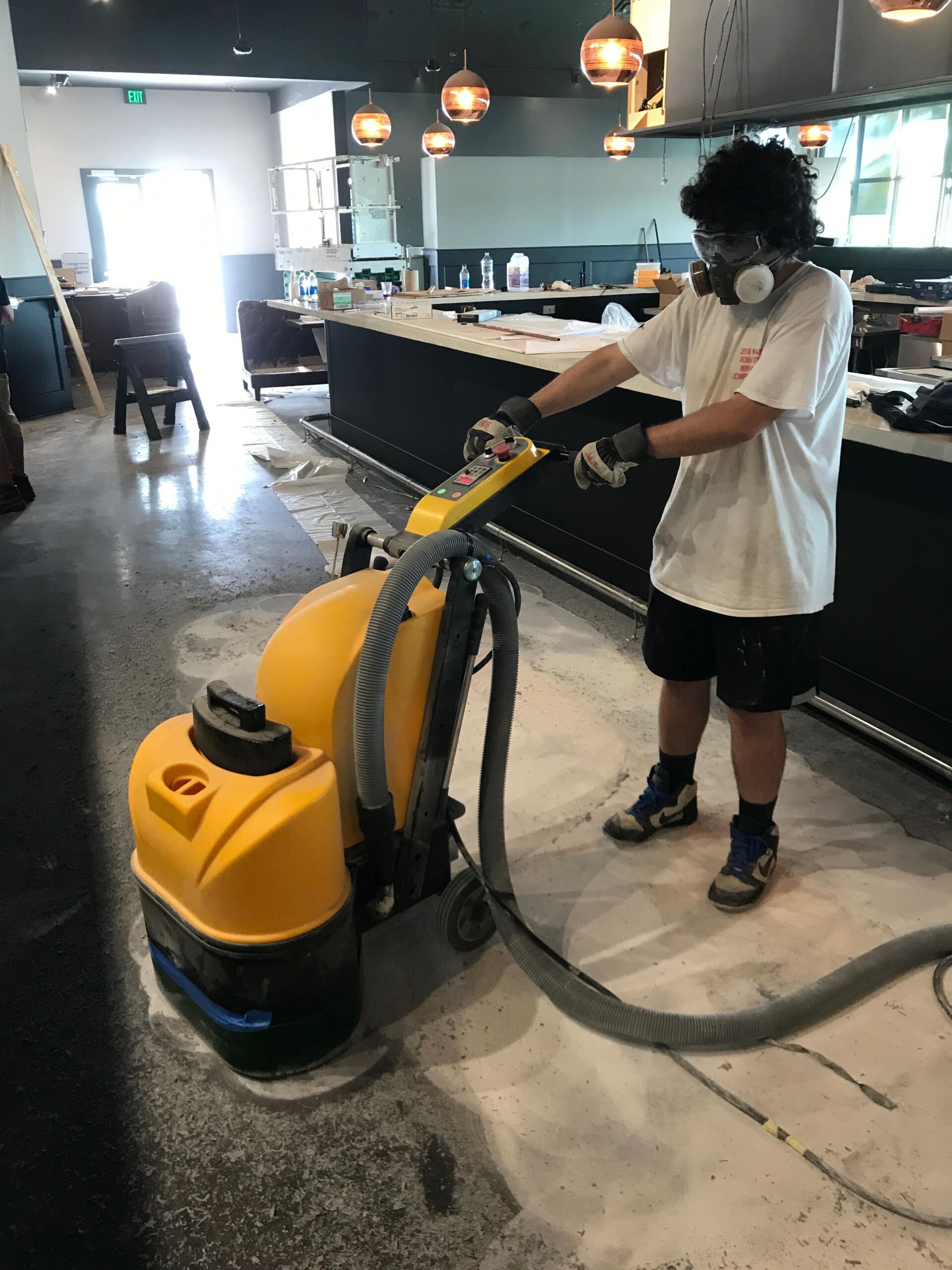 A man wearing a gas mask is using a yellow vacuum cleaner on a concrete floor.