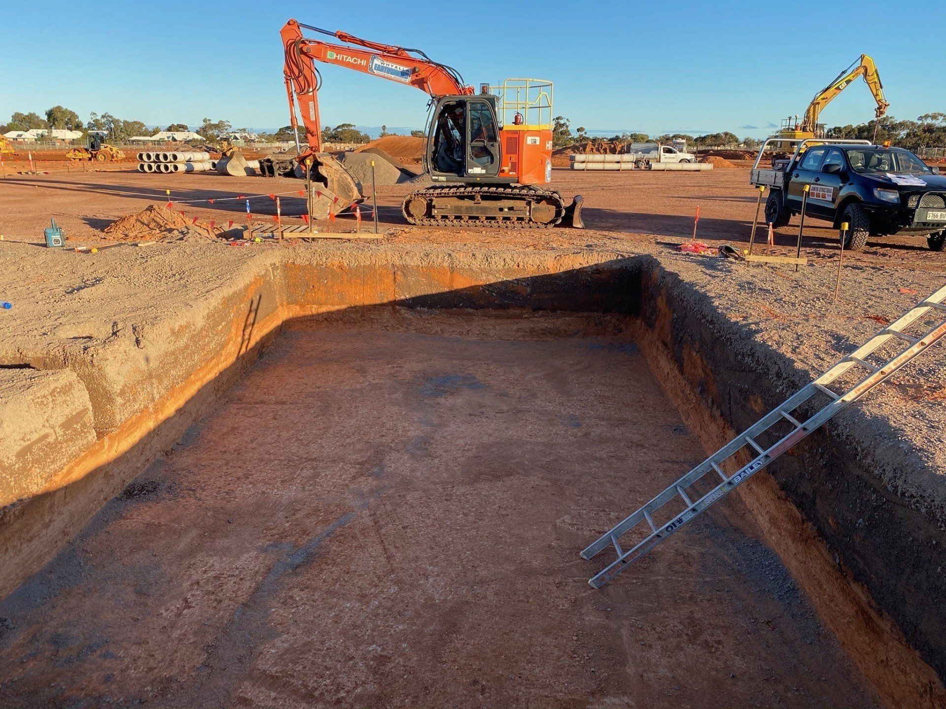 Construction site: Excavator in a large rectangular pit. A ladder is in the pit. Red dirt and blue sky.
