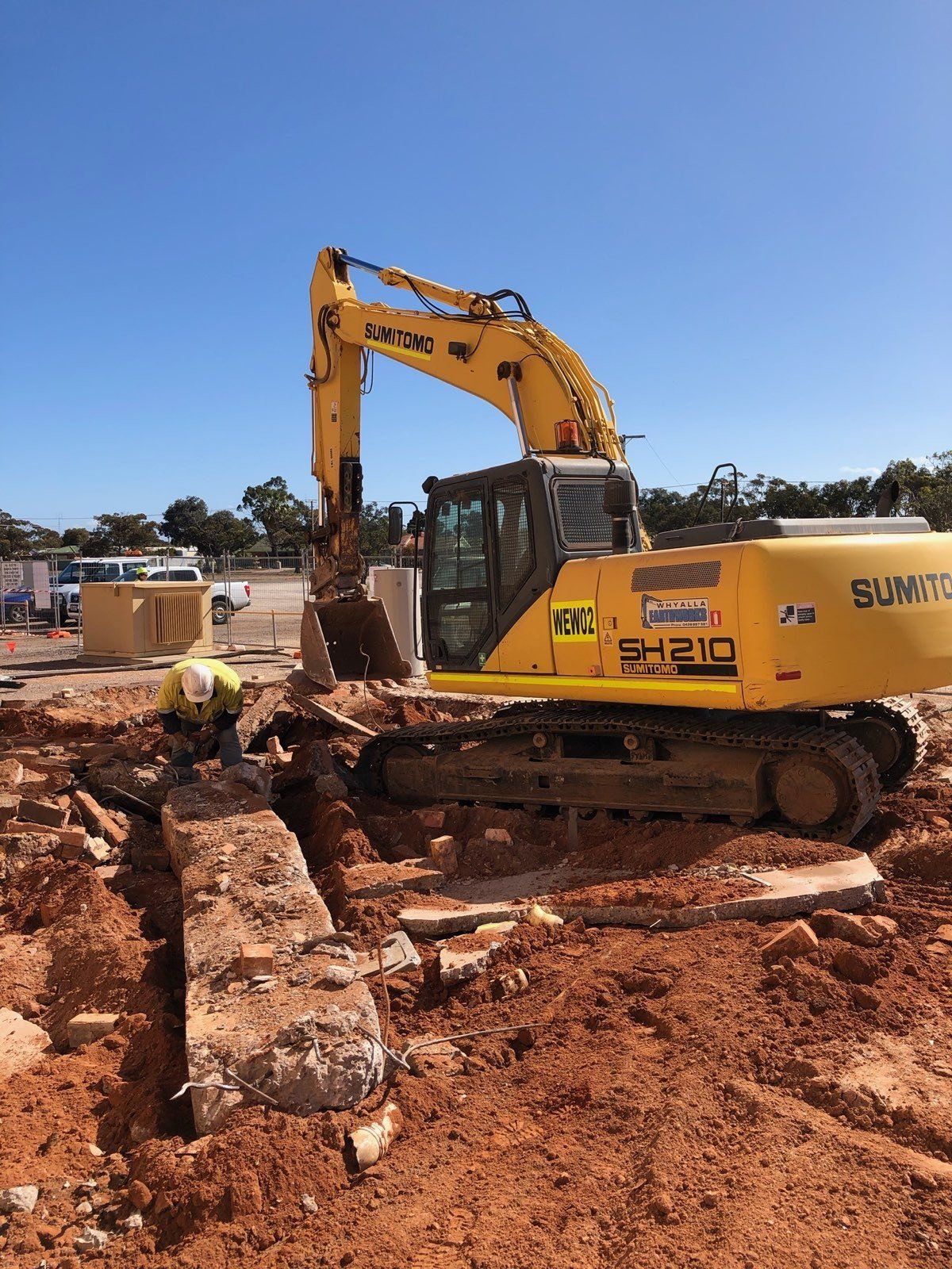 Yellow excavator demolishing concrete structure; construction worker nearby. Sunny day, outdoor setting.