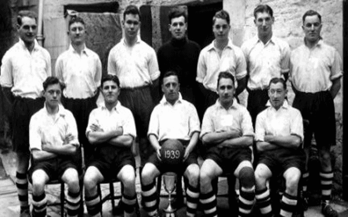 Black and white team photo of a soccer team, likely from 1939. Eleven men in white shirts and dark shorts pose with a ball.