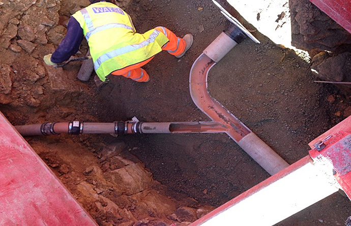 Worker in yellow vest repairing pipes in a ditch, using tools. Earth and machinery visible.