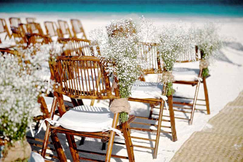 Una fila de sillas plegables de madera decoradas con flores de aliento de bebé en una playa.