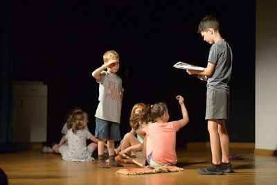 A boy is reading a book to a group of children on a stage.