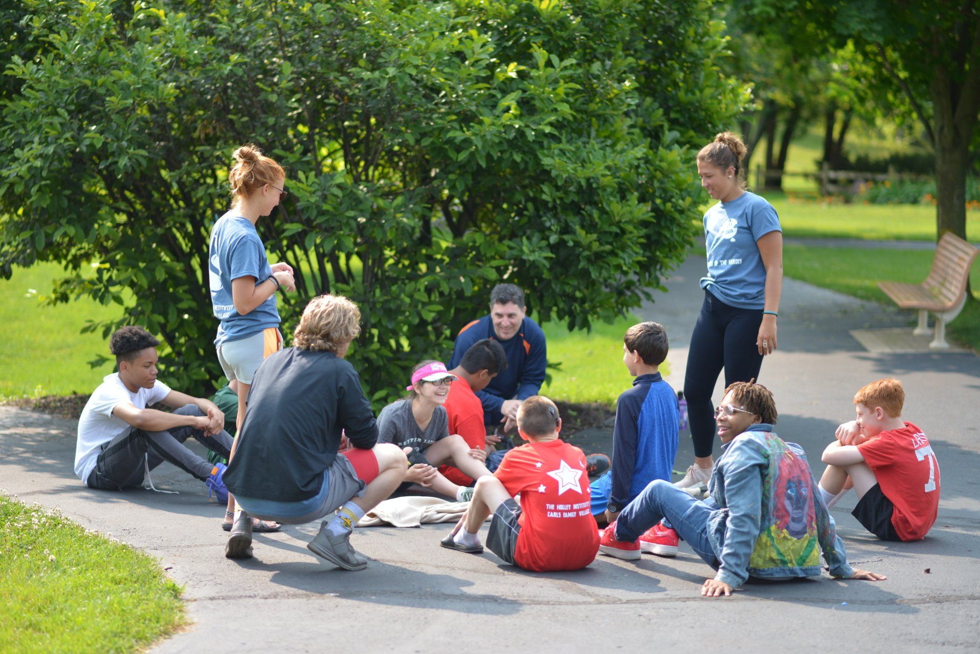 A group of children are sitting in a circle on the sidewalk in a park.