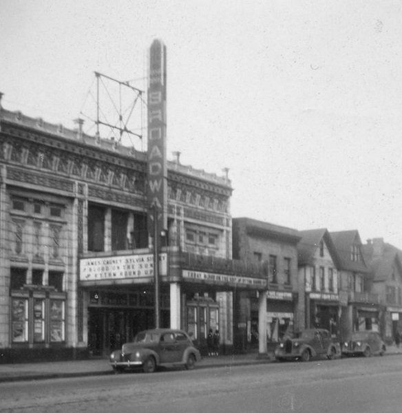 A black-and-white street-level view of the Broadway Theater with cars parked in front under its vertical marquee.