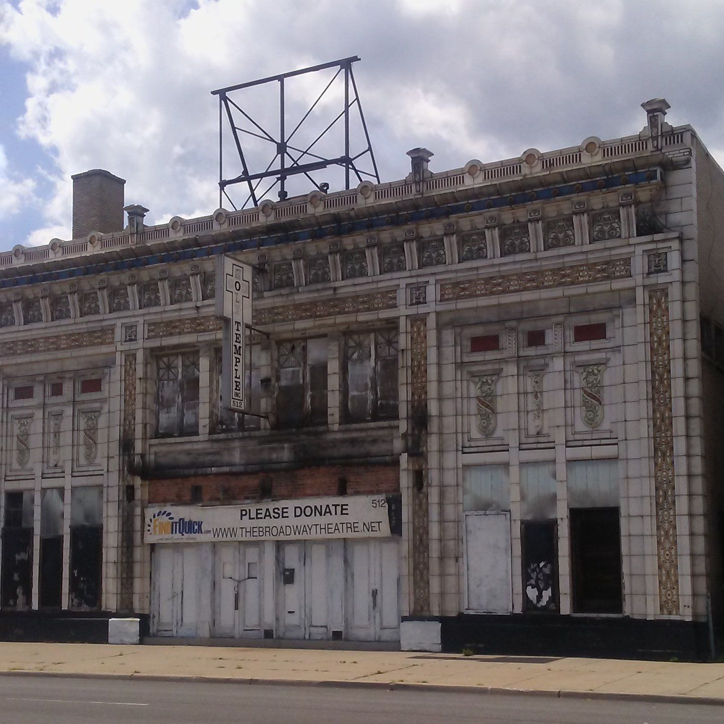 An ornate, aged two-story building with boarded-up ground-floor windows and a donation sign under a metal rooftop frame.