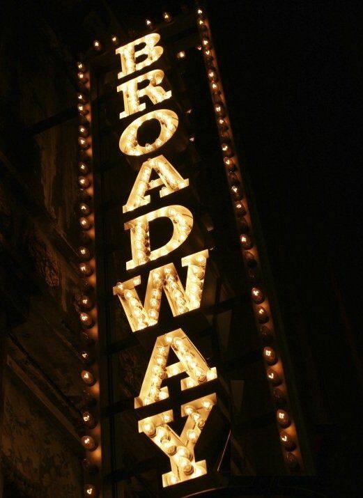 A vertical, illuminated Broadway sign featuring bright marquee lights against a dark background.