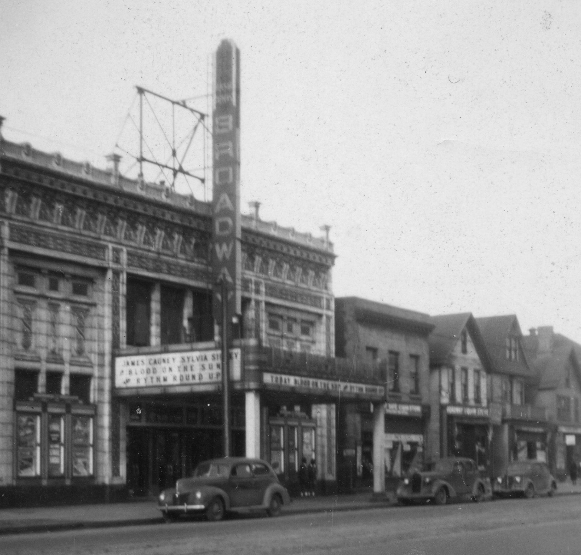 Black-and-white street-level photo of the Broadway Theater building with a vertical sign and parked vintage cars.
