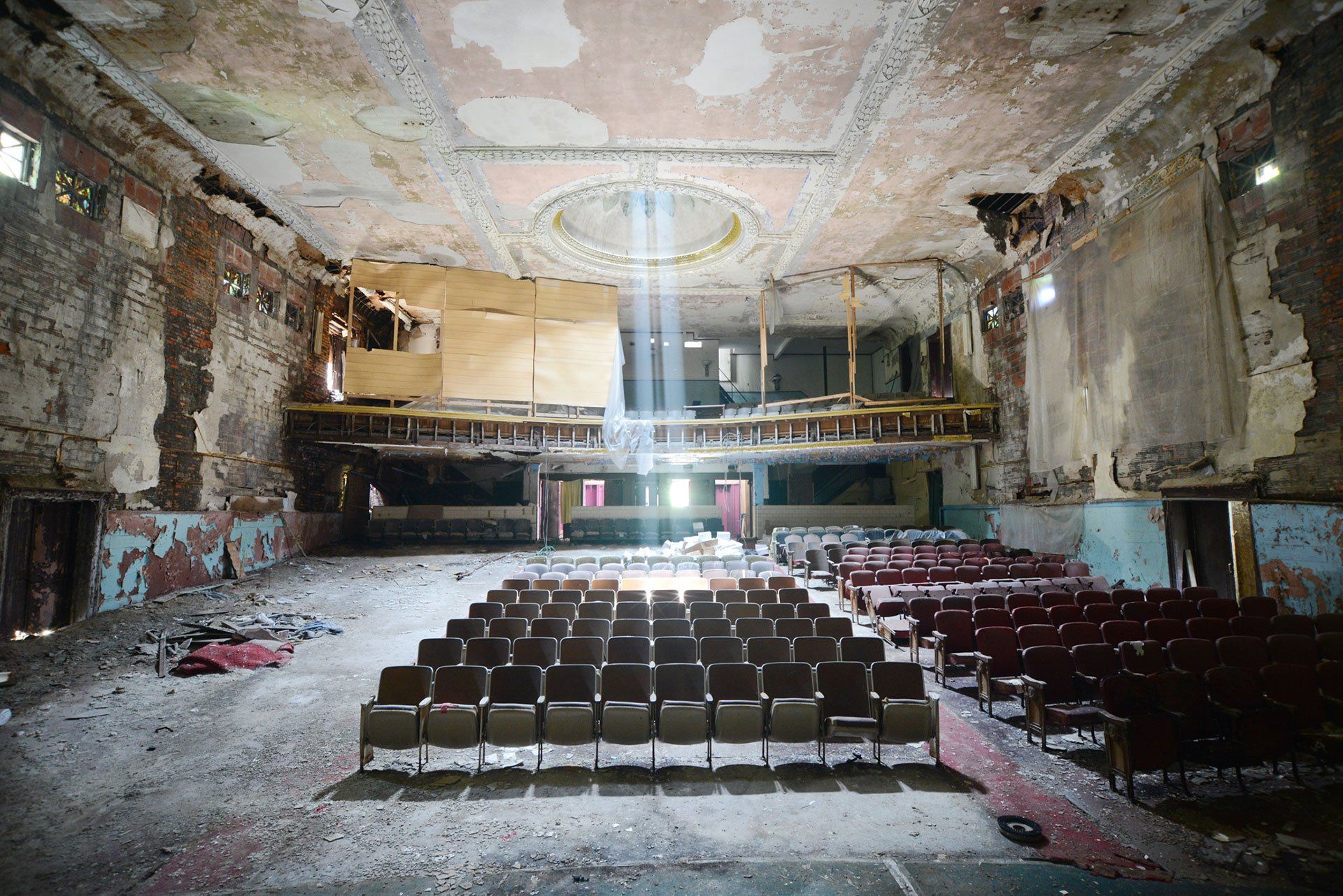 An abandoned, dilapidated theater with rows of empty seats under a sunlit ceiling opening.