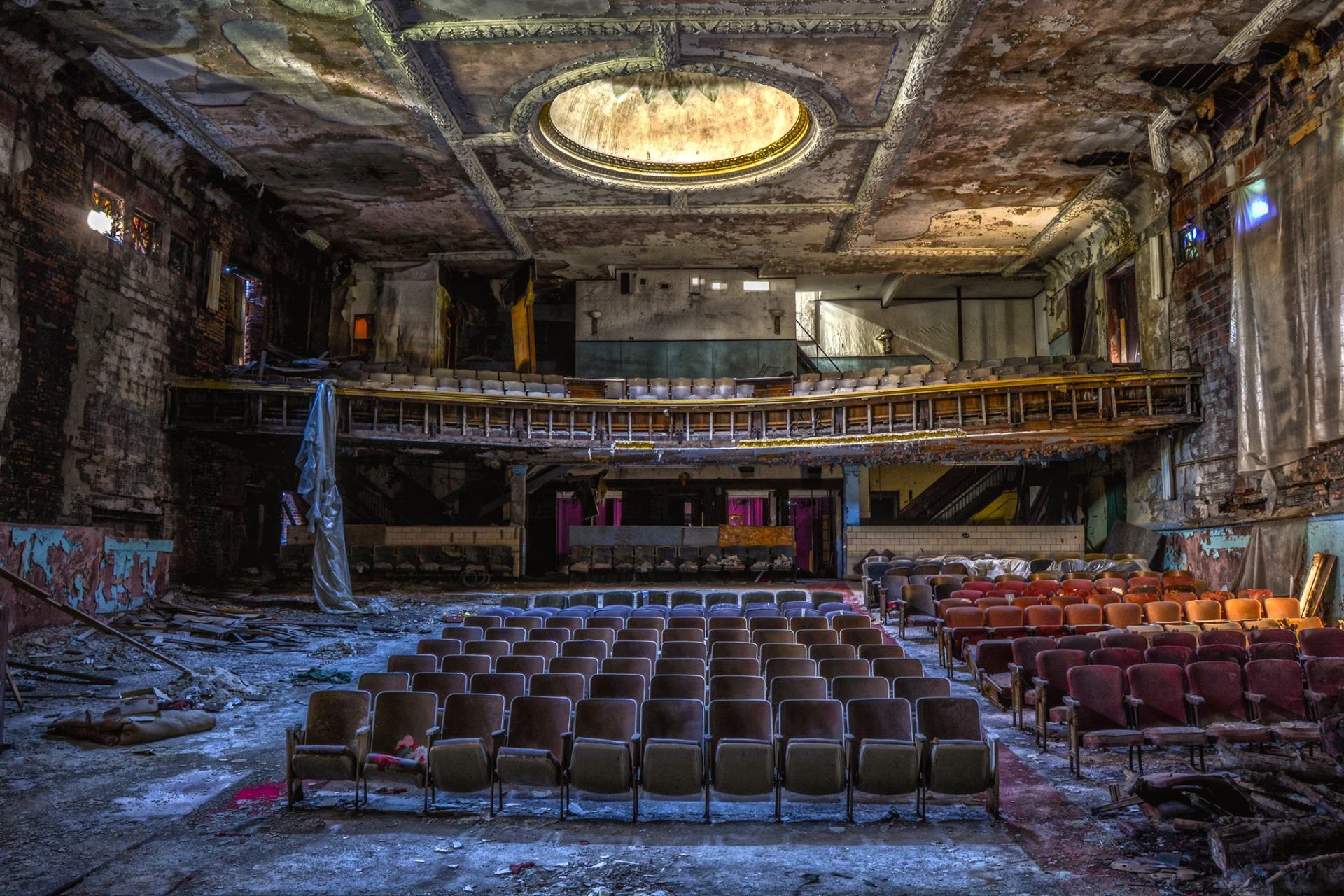 Dilapidated theater interior with rows of empty seats facing a stage under a decaying domed ceiling.