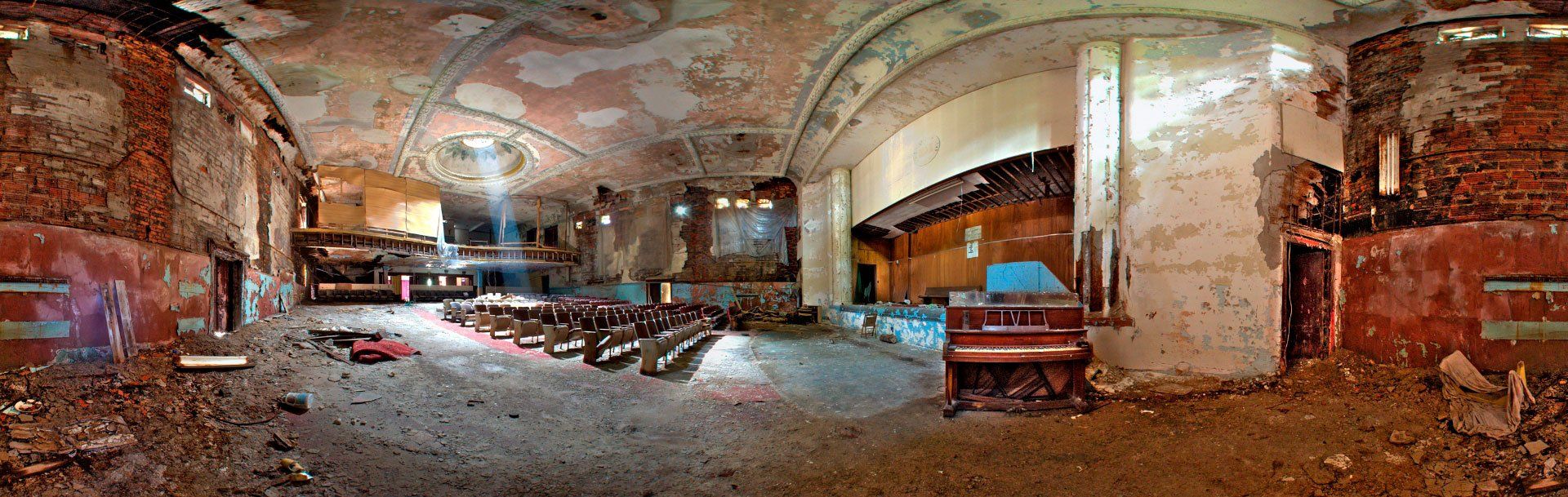 A panoramic view of a decayed, abandoned theater interior with peeling walls, rubble, and rows of empty, damaged seats.