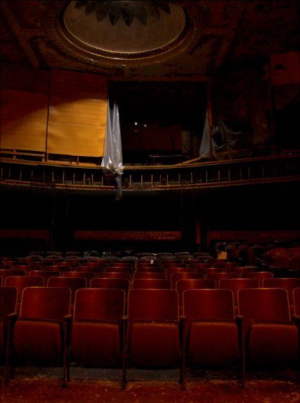 Rows of empty red velvet seats in a dim, decaying theater with a circular ceiling ornament and a dark balcony.