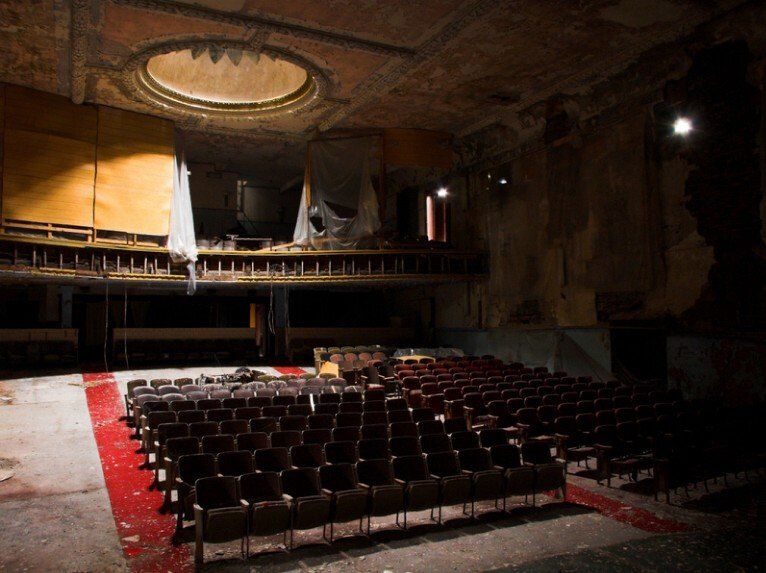 An abandoned, dimly lit theater features rows of empty, dark seats and a dilapidated balcony under a circular ceiling dome.