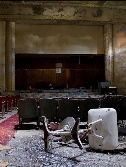 An abandoned theater with rows of dark seats, a stage in the background, and a white chair lying on a debris-covered floor.