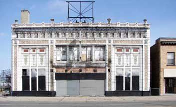 A two-story commercial building with an ornate white facade, large windows, and a metal sign frame on the flat roof.