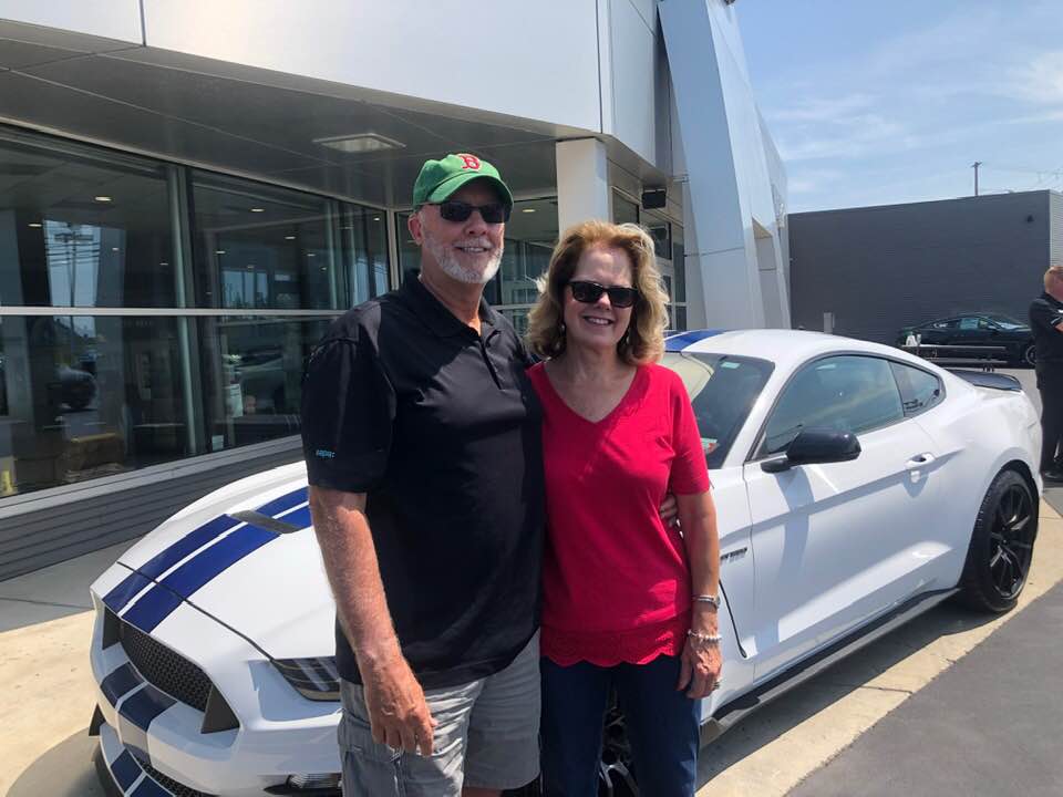 A smiling couple stands in front of a white Ford Mustang with blue racing stripes outside a car dealership.