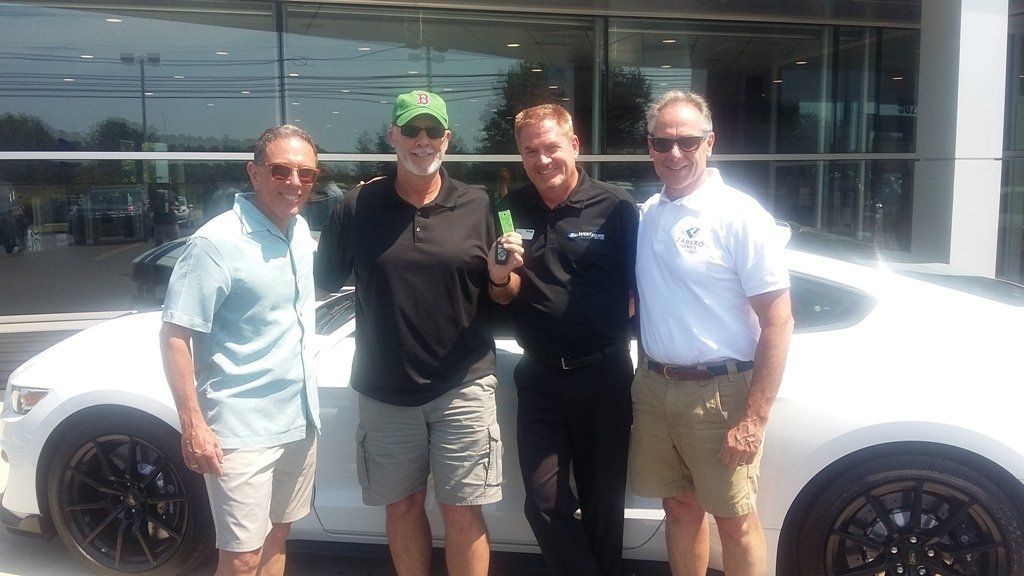 Four men smiling in front of a white sports car outside a dealership, one holding a set of keys.
