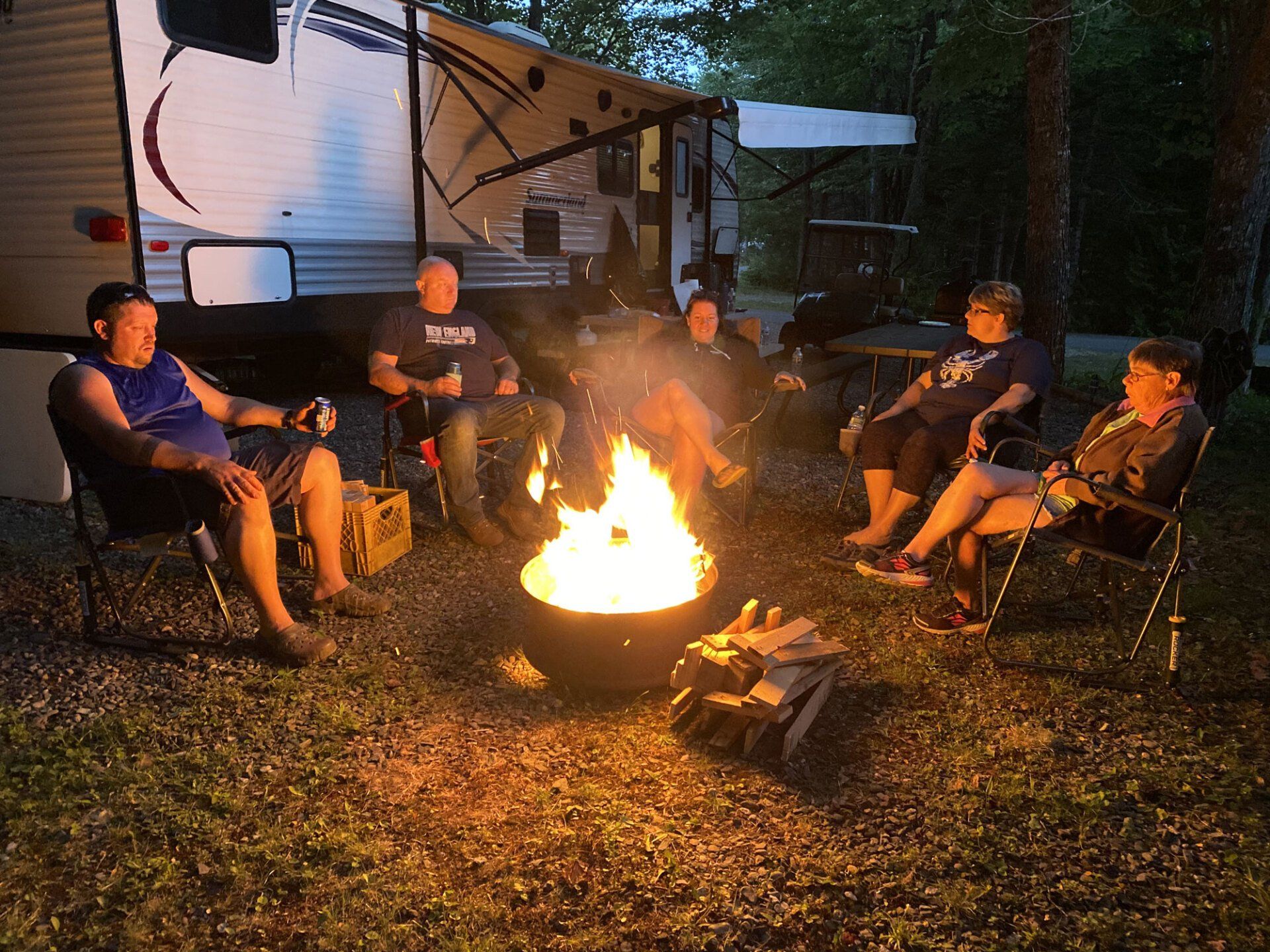 People sitting around a glowing campfire beside an RV at dusk.