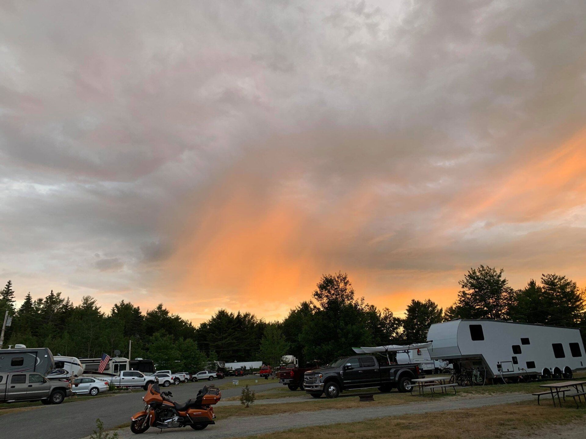 Orange sunset over a campground with RVs, trucks, and an ATV near a tree line under cloudy sky